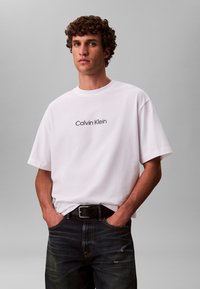 Hvit bomulls-t-skjorte med korte ermer, med en svart "Calvin Klein"-logo over brystet. Kombinert med mørkeblå jeans og et svart belte.