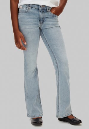 Osoba w jasnoniebieskich dżinsach z rozszerzanymi nogawkami i czarnych płaskich butach, jedna ręka w kieszeni, druga swobodnie opuszczona wzdłuż ciała.