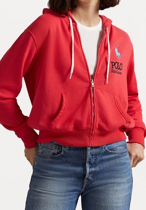 Osoba nosząca czerwoną bluzę z kapturem na zamek z białymi sznurkami i logo Polo Ralph Lauren, w połączeniu z niebieskimi dżinsami i białą koszulą pod spodem.
