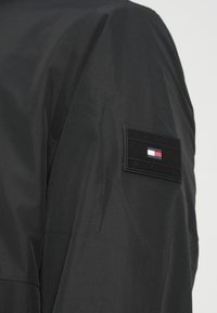 Μαύρο μπουφάν από νάιλον με υφή υφάσματος, με ορθογώνιο μαύρο μπάλωμα με την επιγραφή "TOMMY HILFIGER" και ένα μικρό κόκκινο, λευκό και μπλε λογότυπο.