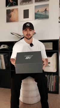 Egy szürke New Balance cipősdoboz, amelyet egy személy tart, fehér logóval. A háttérben polcok találhatók, amelyeken lemezek és sapkák láthatók. A személy fehér pólót visel.