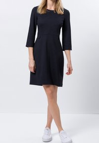 Marineblaues Kleid mit tailliertem Oberteil, Dreiviertelärmeln und Taschendetails. Glatte Textur, knielang, kombiniert mit weißen Sneakers.