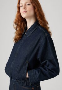 Veste en denim bleu foncé avec col, taille élastique et poches latérales. Dotée d'une fermeture éclair à l'avant et de détails de couture contrastants.