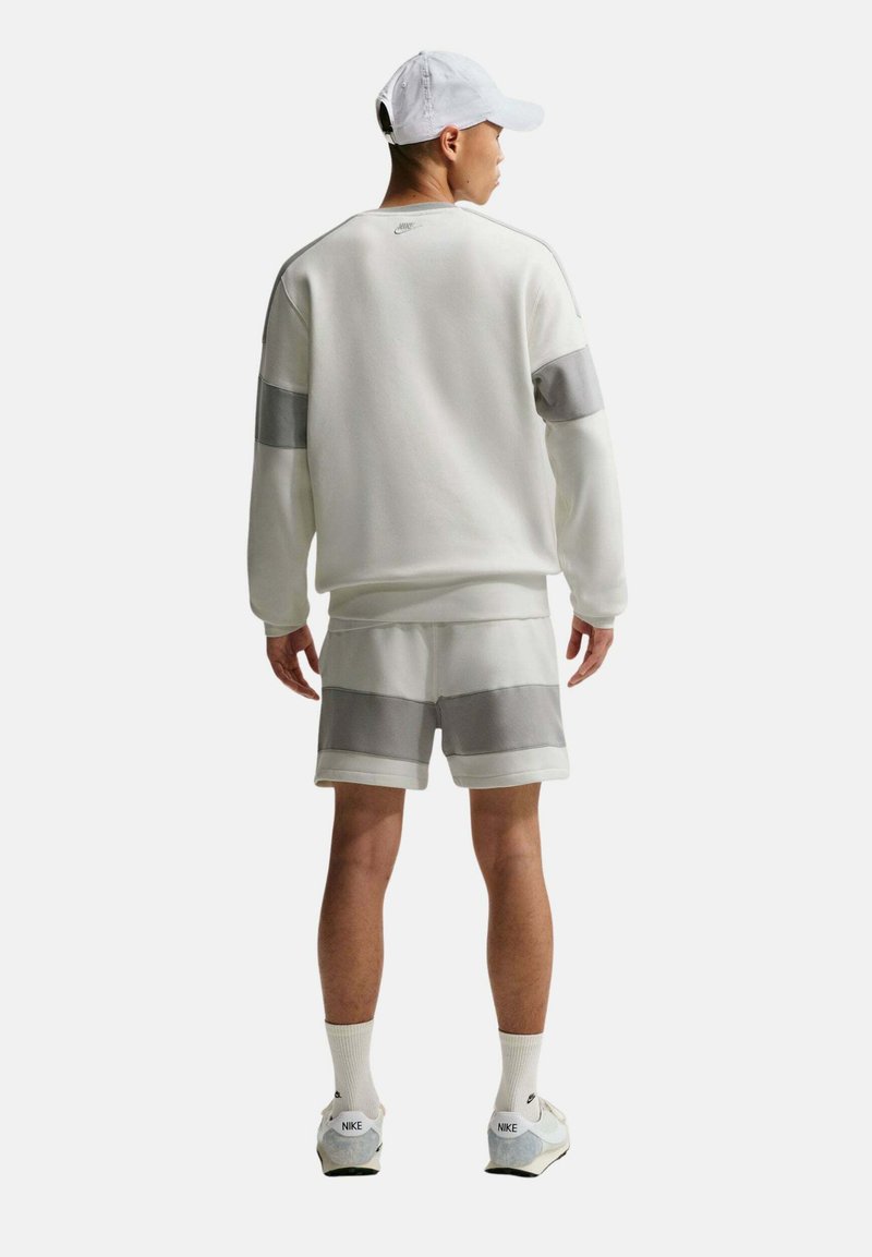 Hombre con gorra blanca, sudadera blanca de manga larga y pantalones cortos a juego con bandas grises, de pie de espaldas sobre fondo blanco.