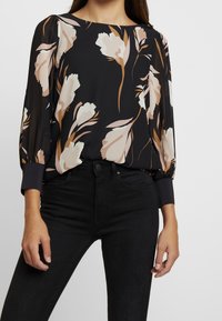 Donna che indossa una blusa nera a fiori con fiori beige e marroni e jeans neri aderenti, in piedi contro uno sfondo semplice.