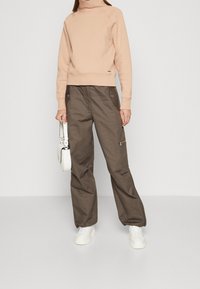 Sudadera de cuello alto en rosa claro con puños acanalados, combinada con pantalones cargo sueltos de color verde oliva que cuentan con bolsillos laterales y cremalleras. Zapatillas blancas completan el conjunto.