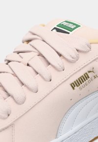 Gros plan sur une sneaker Puma Suede rose clair avec des lacets épais, un logo doré et des détails de surpiqûre blanche sur la semelle.