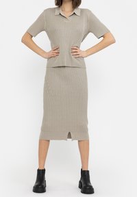 Beige geribbelde trui met korte mouwen en polokraag, gecombineerd met een bijpassende rok met een split aan de voorkant. Zwarte enkellaarsjes maken de look compleet.