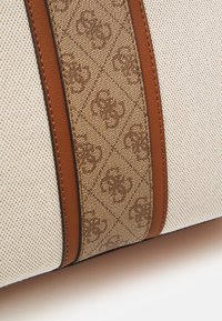 Sac beige texturé avec des accents en cuir brun et un détail à motifs. Présente une bande beige avec un design monogramme. Forme structurée.