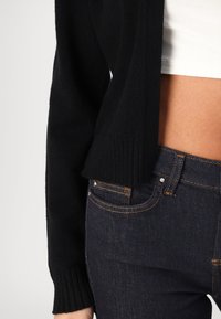 Nahaufnahme einer Person in dunkelblauen Denim-Jeans, einem kurz geschnittenen schwarzen Strickpullover und einem weißen Oberteil, das den Bauch und die Hand an der Seite zeigt.