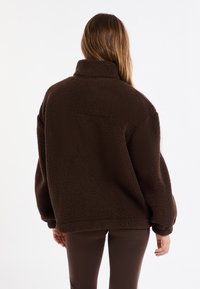 Brązowa polarowa kurtka z wysokim kołnierzem, luźnym krojem i teksturowanym materiałem, noszona na ciemnobrązowych spodniach.