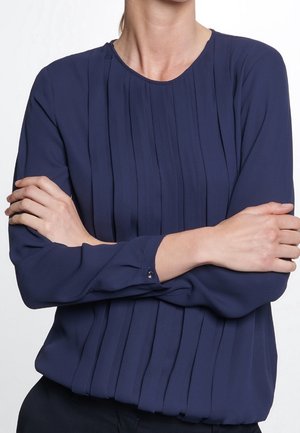 Person iført en langærmet marineblå bluse med lodrette plisseringer, armene krydset foran en ensartet hvid baggrund.