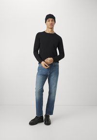 Un top negru cu mâneci lungi, blugi din denim albastru și pantofi negri cu șireturi. Persoana poartă o căciulă închisă și stă pe un fundal gri deschis.