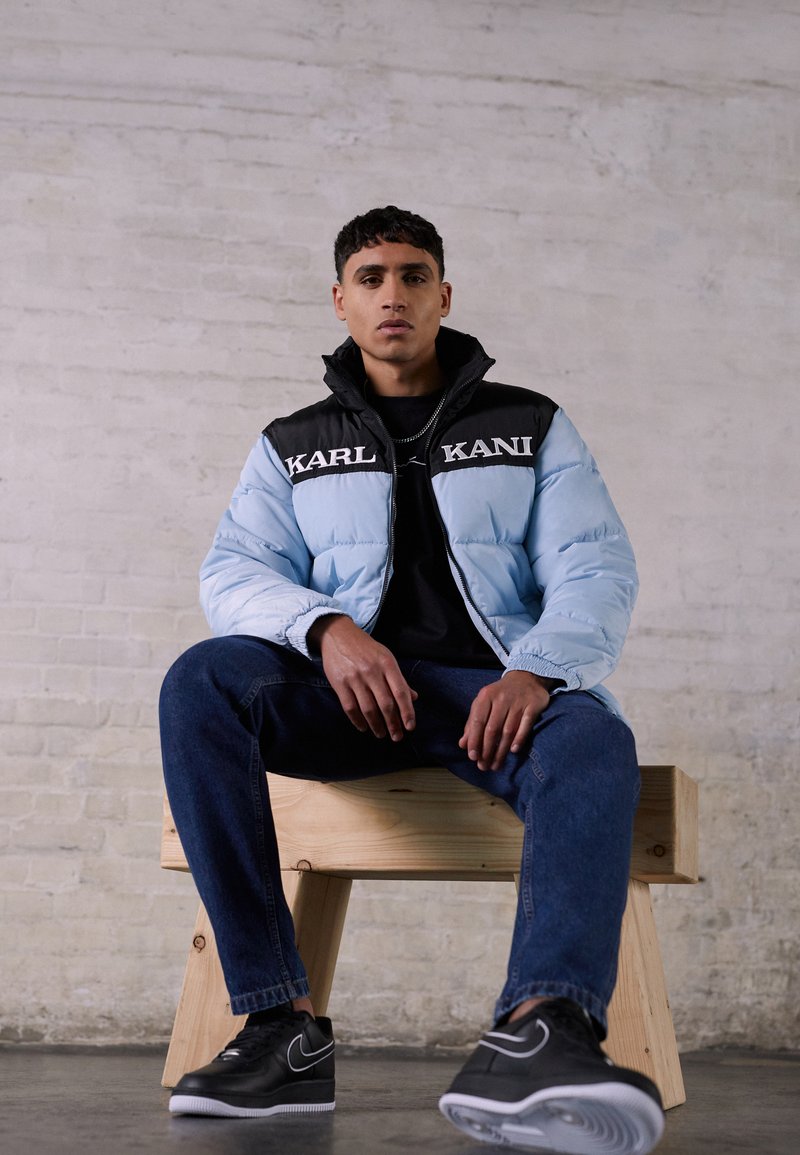 Lyseblå dunjakke med svarte detaljer og logo, iført over en svart skjorte, kombinert med blå jeans og svarte sneakers, sittende på en trebenk.