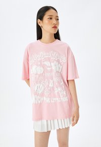 Pink oversized T-shirt lavet af bomuld med hvide grafiske tryk, herunder blomster, sol og motiverende tekst. Afslappet pasform med ribbet hals.