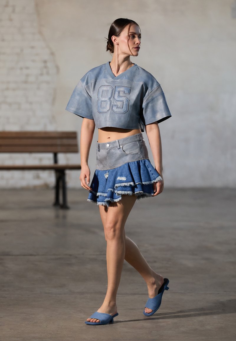 Camisa de cuero azul corta con el número 85, falda de mezclilla acampanada con dobladillo deshilachado y sandalias de tacón abierto azul a juego.