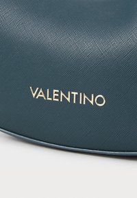 Mėlynos spalvos tekstūruota rankinė, su kreivės forma ir ryškiai matomu auksiniu "VALENTINO" logotipu. Medžiaga turi švelnų grūdėtumą.