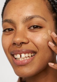 Modella con un anello al naso in oro che applica un fondotinta leggero sulla guancia. Ha ricci corti, pelle liscia e smalto per unghie neutro.