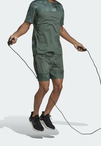 Person som hoppar hopprep i grön träningsklädsel och svarta löparskor mot en ljus, slät bakgrund.