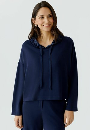 Femme aux cheveux mi-longs et foncés portant des boucles d'oreilles argentées en forme d'anneaux et un sweat à capuche bleu marine avec cordons, souriant légèrement, les mains détendues.