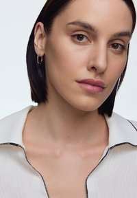 Biała koszula o teksturowanej powierzchni z czarnymi wykończeniami, posiadająca luźny kołnierzyk. Modelka ma krótkie ciemne włosy i małe srebrne kolczyki w kształcie kół. Naturalny makijaż.