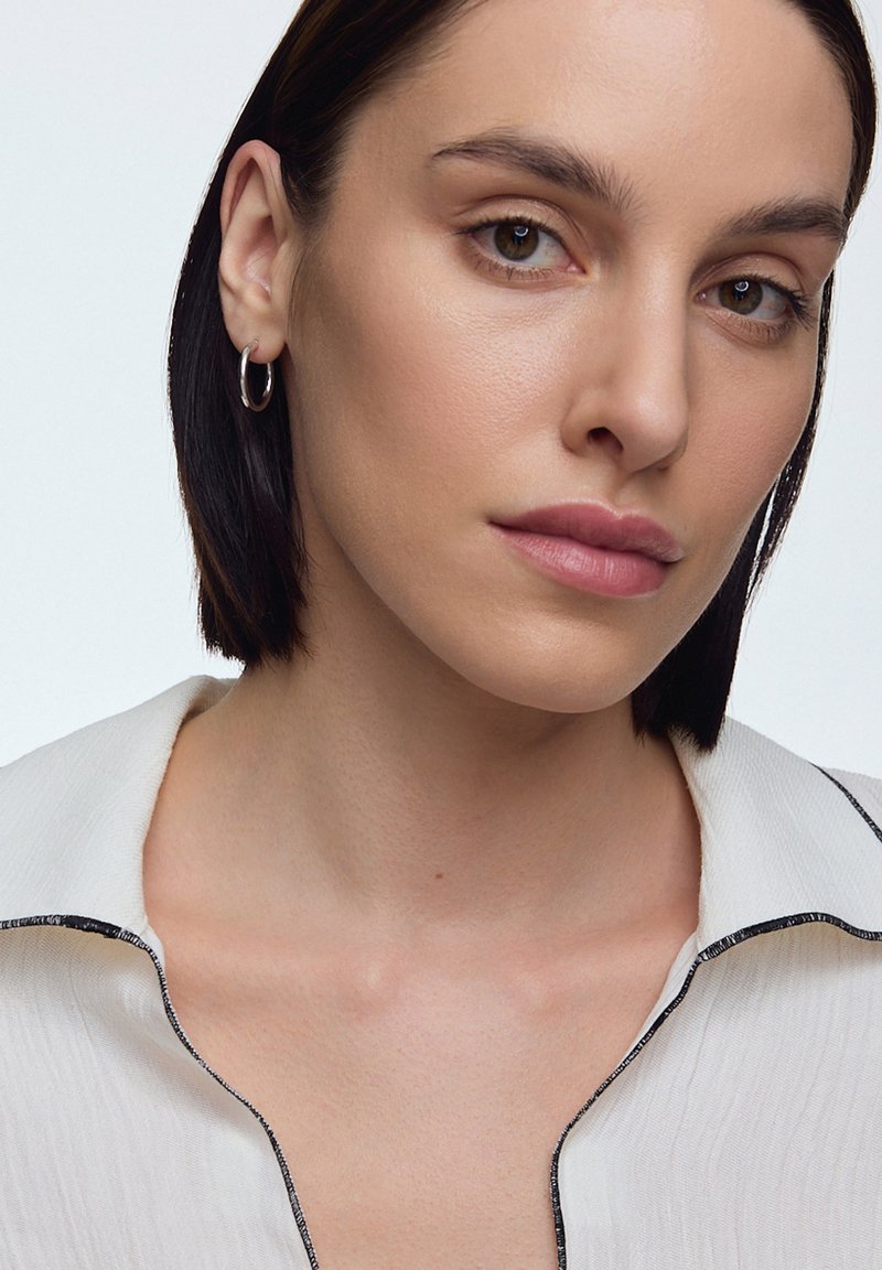 Biała koszula o teksturowanej powierzchni z czarnymi wykończeniami, posiadająca luźny kołnierzyk. Modelka ma krótkie ciemne włosy i małe srebrne kolczyki w kształcie kół. Naturalny makijaż.