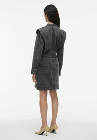 Femme se tenant debout portant une robe en denim structurée gris foncé avec des épaulettes et des chaussures à talons beiges, de dos sur un fond blanc.