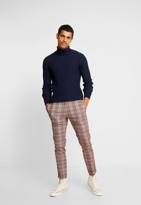 Un pull à col roulé côtelé bleu marine associé à un pantalon à carreaux marron. La tenue présente une silhouette ajustée et des baskets montantes de couleur crème.