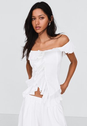 Donna con capelli lunghi e scuri che indossa un top bianco con spalle scoperte e volant e una gonna abbinata, in piedi con le mani dietro la schiena.