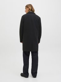 Palton negru cu un design croit, fante unică la spate și rever cu câte o crestătură. Asortat cu pantaloni din denim închis la culoare și pantofi negri.