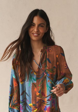 Žena s dlhými tmavými vlasmi sa usmieva, nosí farebnú kvetovanú blúzku s modrými a hnedými vzormi, drží okuliare za ručnú časť.