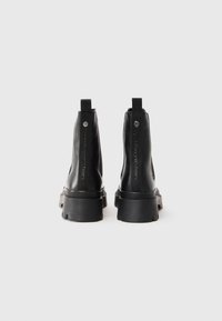 Een paar zwarte enkelboots met een chunky zool, met de hielen naar achteren gericht, voorzien van trektabs en geperste branding op de achterste band.