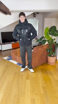 Μαύρο μπουφάν με γυαλιστερό πάνελ, ριγωτό μαύρο παντελόνι και λευκά αθλητικά παπούτσια. Καφέ καπιτονέ καναπές και πράσινο φυτό στο φόντο.