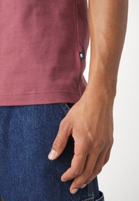 Rechterhand van een persoon die een bordeauxrood shirt en blauwe jeans draagt, met een klein zwart stoffen label op de zoom van het shirt.