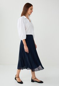 Blusa bianca, ampia e svasata con maniche a palloncino abbinata a una gonna plissettata lunga fino alla caviglia di un blu navy scuro. Ballerine nere con cinturini alla caviglia.
