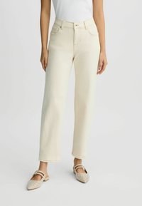 Femme portant un jean crème taille haute, coupe droite, et des ballerines beiges à clous pointues, se tenant contre un fond uni clair.