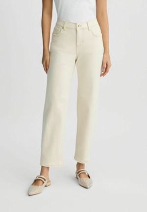 Donna che indossa pantaloni in jeans a vita alta, modello dritto, di colore crema e ballerine beige con borchie, in piedi davanti a uno sfondo chiaro e semplice.