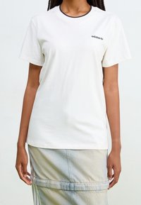 Valkoinen puuvillainen lyhythihainen t-paita, pyöreä pääntie, jossa tumma reuna, ja pieni musta Adidas-logo vasemmassa rinnassa.