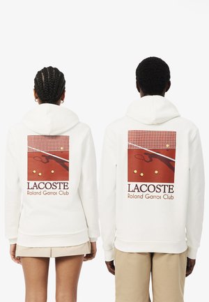 Deux personnes portant des sweat-shirts blancs à capuche avec un graphisme rouge représentant un court de tennis, des balles de tennis et le texte « Lacoste Roland Garros Club » dans le dos.