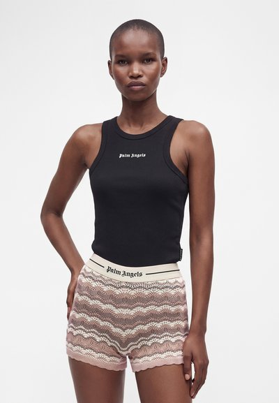 Femme portant un top noir sans manches Palm Angels et un short en maille rose à motifs avec une ceinture à logo Palm Angels, debout devant un fond blanc.