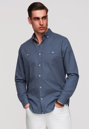 Junger Mann mit gestyltem Haar, der ein blaues Hemd mit Brusttaschen und weiße Hosen trägt, die Hände in den Taschen, schaut zur Seite.