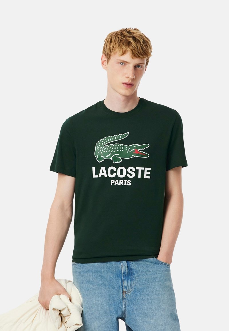 Camiseta negra de algodón con un gráfico de cocodrilo verde y el texto "LACOSTE PARIS". El modelo sostiene una prenda blanca en la mano.