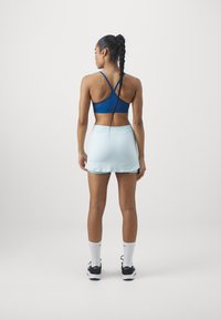 Mujer con el cabello trenzado largo de espaldas, vistiendo un sujetador deportivo azul, una falda deportiva azul clara, calcetines blancos y zapatillas negras.
