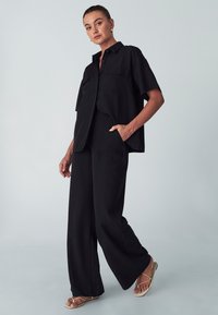 Černý dvoudílný outfit skládající se z košile s krátkým rukávem na knoflíky a kalhot s širokými nohavicemi. Vyrobeno z hladké tkaniny s uvolněným střihem a předními kapsami.