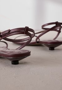 Stilvolle burgunderrote Sandalen mit dünnen Lederbändern, minimalistischem Design und niedrigem, kegelstumpfförmigem Absatz mit runden schwarzen Akzenten.