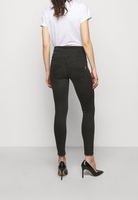 Jeans ajustados de tiro alto en gris oscuro con una textura suave, dos bolsillos traseros con costuras decorativas y un corte slim. Combinados con tacones negros.
