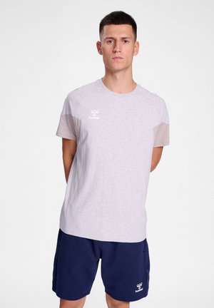 Man staat met zijn handen achter zijn rug, draagt een lichtgrijze Hummel t-shirt en marineblauwe shorts tegen een effen witte achtergrond.
