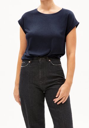 Kvinde iført en mørkeblå kortærmet top stoppet ned i højtaljede sorte jeans, der står mod en ensfarvet hvid baggrund.