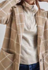 Cárdigan de cuadro beige hecho de material suave, con grandes bolsillos, superpuesto a un suéter de canalé crema con cuello de media cremallera.