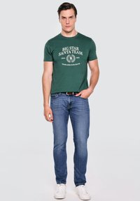 Zelené bavlněné tričko s bílým textovým designem, spárované s modrými džíny a bílými teniskami. Model stojí na neutrálním pozadí.